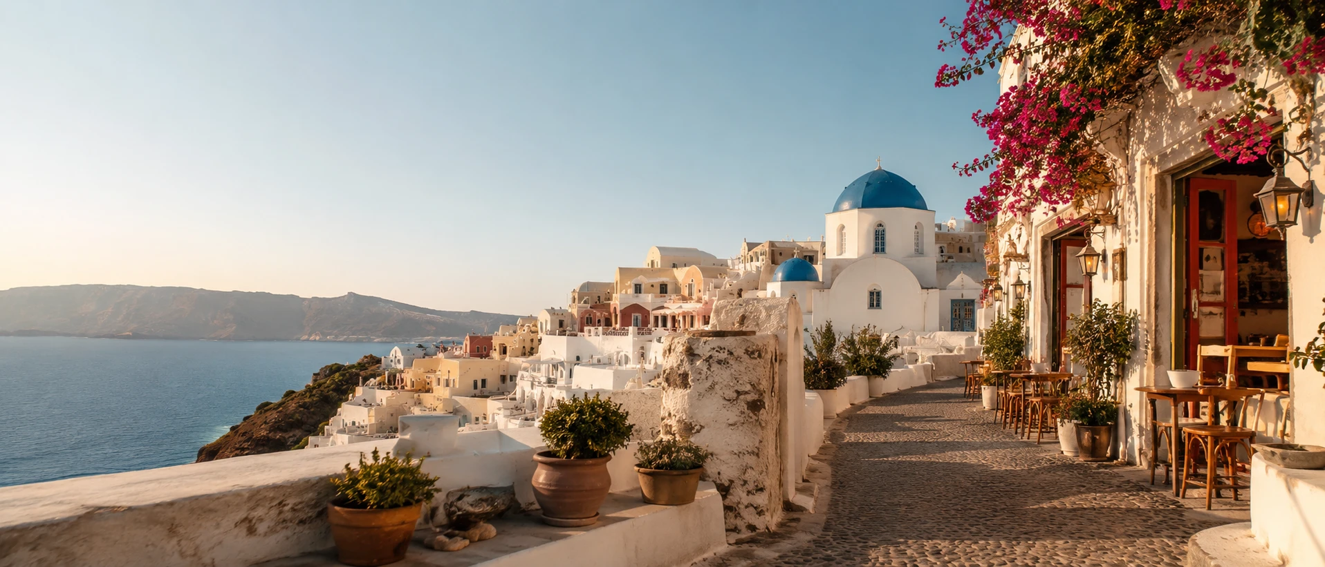 A cobbled clifftop lane in Oia, Santorini, past blue-domed churches and a bougainvillea-draped café at golden hour